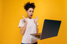 Stunned Shocked African American Girl, In A T-shirt, With Curly Hair, Looks In Surprise At The Laptop Screen, Gesticulates With Her Hands, Saw The Unexpected News, Stands On Isolated Orange Background