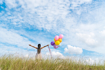 Wall Mural - Cheerful cute girl holding balloons running on green meadow white cloud and blue sky with happiness. Hands holding vibrant air balloons play on birthday party happy times summer on sunlight outdoor