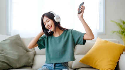 Young asian woman listening to music on couch in living room at home. Happy asia female using mobile smartphone, wearing headset and sitting on sofa