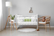 Interior of light nursery with baby crib, lamp, table and armchair