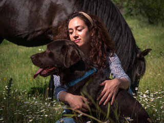 Horizontal view of female with her chocolate labrador retriever in the mountains in spring meadow and black horse in the background.