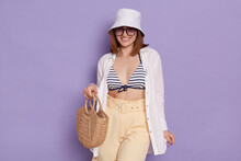 Indoor Shot Of Smiling Relaxed Woman Wearing Swimsuit, Shirt, Shorts And Panama Posing Isolated Over Purple Background, Standing With Straw Bag And Eyeglasses, Being Ready Going To The Beach.