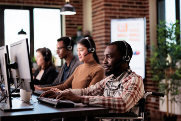 Wall Mural - African american helpline operator with chronic impairment working at call center client care. Man wheelchair user giving telework assistance at customer service in disability friendly office.