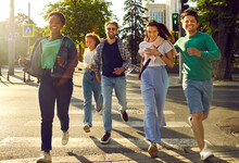 Bunch Of Happy, Carefree Friends Quickly Crossing The Street. Group Of Joyful Young Multiracial People In Casual Clothes Running And Having Fun During Their Evening Hang Out In A Modern European City