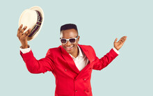 Happy African Man In Funky Outfit Welcomes You At Party. Cheerful Tanzanian Gentleman In Stylish Red Suit Standing Isolated On Blue Background Takes Off His Hat, Smiles And Makes Bow. Fashion Concept