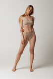 Full-length portrait of beautiful young woman with sportive muscular body posing in beige cotton underwear isolated over grey studio background