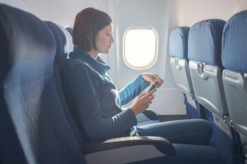 Wall Mural - Young beautiful woman sitting at window of plane during the flight. reading a book on a smartphone, playing apps, writing notes