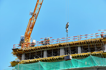 A yellow tower crane lifts a load on a building construction site with a green mesh against a clear blue sky