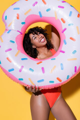 Wall Mural - cheerful african american woman with curly hair holding inflatable ring on yellow background.