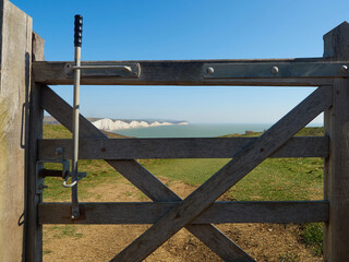 Sunlit white chalk cliffs under summery blue skies near Seaford, seen through the slats of a gate on the hiking trail.