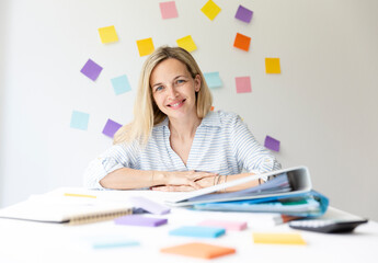 Wall Mural - Portrait of pretty young blonde woman with beautiful laugh sitting at office table in front of white wall with colorful sticky notes