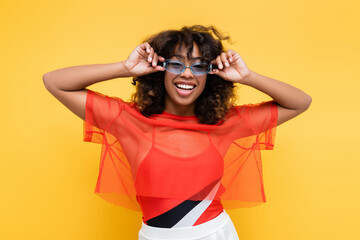 Wall Mural - happy african american woman in trendy summer clothes adjusting sunglasses isolated on yellow.