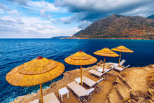 Summer Vacation Destination. Straw Sunshades And Sunbeds On The Empty Pebble Beach With Sea In The Background. Vacation And Tourism Concept. Sunbeds On The Paradise Beach. Umbrellas And Sunbeds.