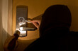 A masked man shining a flashlight on a door that he is trying to open with tools