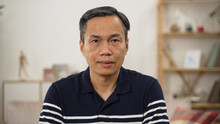 Half Length Portrait Of A Grey Haired Asian Male Pensioner In Casual Wear Looking At Camera With Serious Face Expression On Home Background