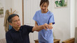© PR Image Factory - Caring female Asian personal nursing aide helping senior stroke patient do rehabilitation training in the living room at home. she lifts his arm slowly