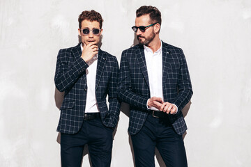 Portrait of two handsome confident stylish hipster lambersexual models. Sexy modern men dressed in black same elegant suit. Fashion male posing in studio near grey wall in sunglasses