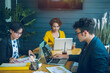 © brain2hands - Confident young businessman sitting at office desk with group of colleagues in office They work by using laptop computers and graphing charts to analyze and plan their business.