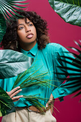 Wall Mural - african american woman in turquoise blouse standing with hand on waist near exotic leaves isolated on pink.