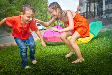 Children Playing With Garden Sprinkler. Brother And Sister Running And Jumping. Summer Outdoor Water Fun In Backyard. Boy And Girl Play With Hose Watering Grass. Kids Run And Splash On Hot Sunny Day