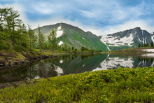 Mountain Lake In Early Spring Under Gloomy Rain Clouds. Coniferous Forest And Rock Ridge On Horizon. Snow On Slopes, On Banks Of River. In Water, Reflection Of Cliffs, Green Grass