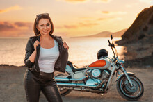 Pretty Smiling Adult Woman In Leather Jacket Posing. Motorbike, Sunset And Ocean On The Background. The Concept Of Motorcyclist Day