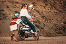 A Beautiful Adult Woman With A High Heels And Leather Pants, Sexy Posing Sitting On Motorcycle. Rear View. Rock On The Background. The Concept Of Motorcyclist Day