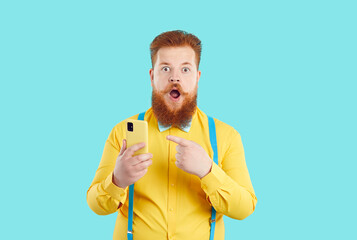 Funny redhead bearded man isolated on blue studio background point at smartphone shocked by unexpected news online. Red-haired guy with mustache surprised with sale or deal on mobile.