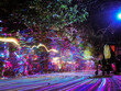© jockermax3d - Bright multi-colored lines glow on the alley in the night park creating a pattern on the defocused background of people walking. Play of light and shadows. Long exposure photo