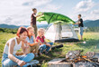 © Soloviova Liudmyla - Three sisters cheerfully laughing and roasting marshmallows and candies on sticks over campfire flame while two brothers set up the green tent. Happy family outdoor picnic camping activities concept.