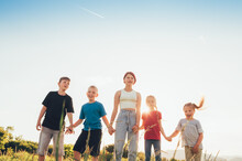 Five Kids Brothers And Sisters Teenagers And Little Kids Walking On The Green Grass Meadow With Evening Sunset Background Light Holding Hands In Hands And Smiling. Happy And Careless Childhood Concept