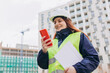 Happy Architect with a blueprints using smartphone at a construction site. Portrait of redhead woman constructor wearing white helmet and safety vest outdoors, technology or builing concept