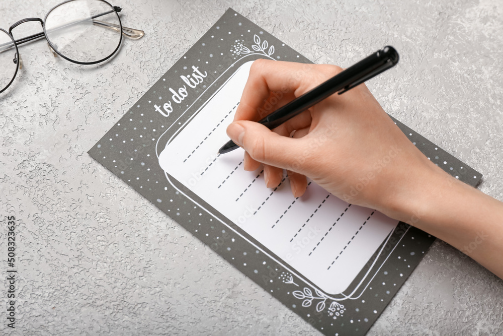 Female hand with to-do list and eyeglasses on grey background, closeup