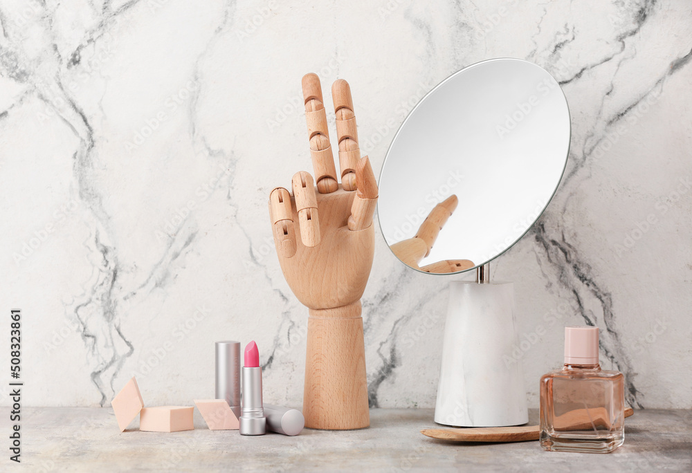 Wooden hand with cosmetic products and mirror on light background