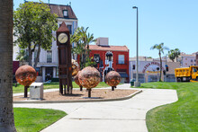A Metal Sculpture Of A Grandfather Clock And Three Large Rusty Balls In The Park Surrounded By Lush Green Palm Trees And Grass And Purple Trees With A Clear Blue Sky At MacArthur Park In Los Angeles