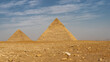 The great pyramids of Cheops and Chephren against the blue sky and clouds. The stones are scattered on the sandy soil of the Giza plateau. Silhouettes of Cairo buildings in the distance. Egypt