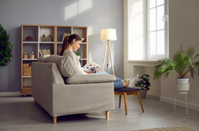 Woman Relaxing On Sofa At Home. Happy Lady Enjoying Couple's Love Story, Wedding Or Honeymoon Photographs In Photo Book While Sitting On Comfortable Couch With Foot Stool In Beautiful Cosy Interior