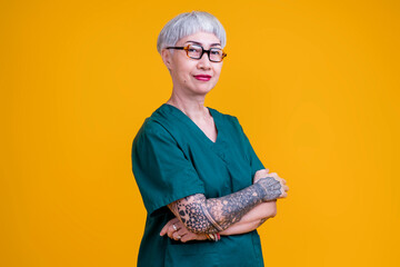 old senior asian female grey hair and tattoo with glasses wear hospital uniform standing with confident and cheerful expression studio shot yellow background