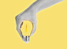 Hand Holding A Light Bulb. The Flame Burning Inside The Bulb. Female Hand Holds Energy Saving Bulb, Isolated On Yellow Background. Photo Manipulation. 3D Illustration.