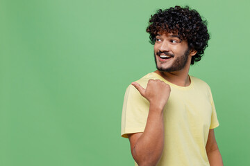 Wall Mural - Young smiling happy Indian man 20s in basic yellow t-shirt point thumb finger aside on workspace area mock up isolated on plain pastel light green background studio portrait. People lifestyle concept.
