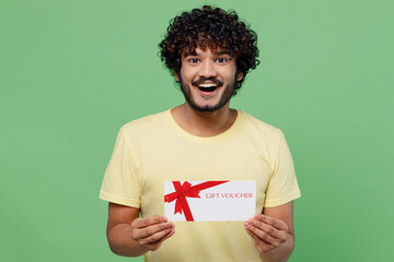 Wall Mural - Young excited happy Indian man 20s in basic yellow t-shirt hold gift certificate coupon voucher card for store isolated on plain pastel light green background studio portrait People lifestyle concept