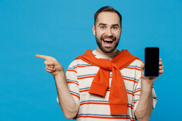 Wall Mural - Young fun surprised amazed man wear orange striped t-shirt hold in hand use mobile cell phone with blank screen workspace point finger aside on area isolated on plain blue background studio portrait