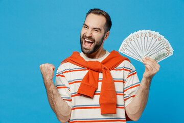 Wall Mural - Young happy excited overjoyed man in orange striped t-shirt holding fan of cash money in dollar banknotes do winner gesture isolated on plain blue background studio portrait. People lifestyle concept.