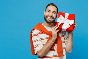 Wall Mural - Young smiling minded pensive happy caucasian man 20s wear orange striped t-shirt hold red present box with gift ribbon bow isolated on plain blue background studio portrait. People lifestyle concept.