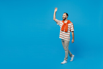 Wall Mural - Full body side profile view young smiling caucasian cheerful man 20s wear orange striped t-shirt walking going waving hand isolated on plain blue background studio portrait. People lifestyle concept.