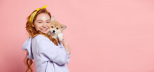 Cheerful Redhead Young Girl With Long Curly Hair Holding Cute Little Puppy Of Corgi Dog Isolated On Pink Background. Concept Of Youth, Beauty, Lifestyle