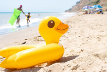 Yellow Inflatable Duck Ring Laying On Sandy Beach Near Blue Wavy Ocean In Sunny Day With People And Sun Umbrellas On Background. Protection Swim Tube For Kid. Summer Travel Vacation Resort Concept