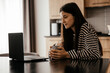 © Максим Галінский - Freelancer woman sits by the table in the home kitchen office, working on laptop. Playful child distracts from work, kid making noise and asking attention from busy mom