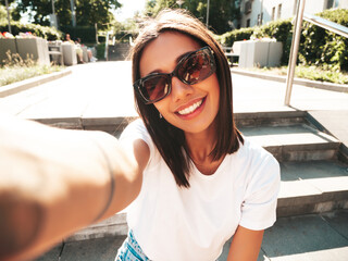 Wall Mural - Portrait of young beautiful smiling hipster woman in trendy summer jeans shorts. Sexy carefree model posing on the street background at sunset. Positive model outdoors.Taking pov selfie