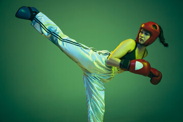 Wall Mural - Leg kick. Flexible female boxer in boxing gloves and helmet practicing isolated on green studio background in neon light. Sport, competition, challenges, results, success concept